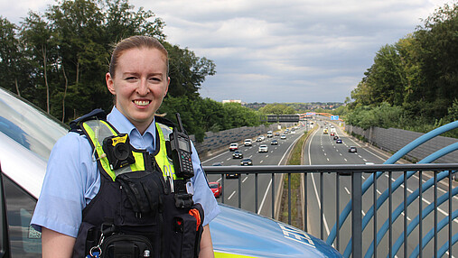 Foto zum Thema "Wie wird man Polizist*in": Zu sehen ist Svenja, die in Uniform vor dem Streifenwagen auf einer Autobahnbrücke steht.