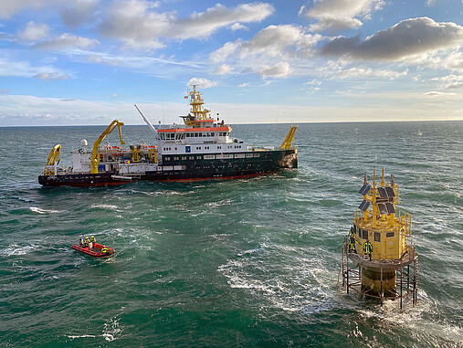 Foto zum Thema "Ausbildung zum Schiffsmechaniker": Zu sehen ist das Schiff "Atair" auf der Nordsee, daneben eine Messboje. Das Team holt Daten ein. Es handelt sich um eine Luftaufnahme.