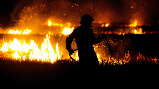 Feuer im Wald - Was es bedeutet, einen Waldbrand zu bekämpfen Foto zum Thema "Feuer um Wald": Zu sehen ist ein Feuerwehrmann, im Hintergrund lodern Flammen im Wald.