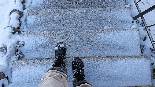 Das Foto zeigt eine zugefrorene Treppe und die Füße einer Person, die diese gerade vorsichtig hinabsteigt.