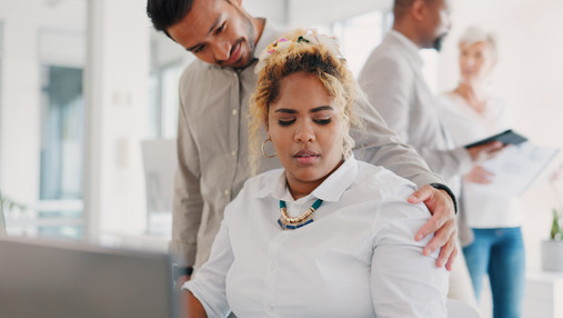 Sexuelle Belästigung am Arbeitsplatz - FAQ für Betroffene Foto zum Thema sexuelle Belästigung am Arbeitsplatz: Zu sehen ist eine Frau, die am Schreibtisch im Büro sitzt, und von einem Mann an der Schulter berührt wird. Ihr Blick verrät, dass es ihr nicht gefällt.