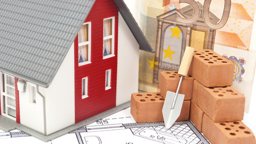 Ein Schritt von großer Tragweite Zu sehen ist ein Modellhaus, daneben ein paar Bausteine, dahinter ein Geldschein. Das Foto ist ein Symbolbild für Immobilienfinanzierung.