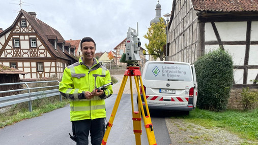 Foto zum Thema "Techniker für Ländliche Entwicklung": Zu sehen ist Andres Greser, der im Job arbeitet. Er steht auf einer Dorfstraße, neben ihm das Messgerät auf einem Stativ.