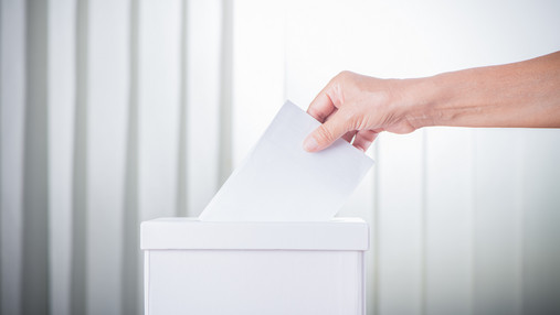 Einmal Wahlhelfer, immer Wahlhelfer - Ehrenamt Foto zum Thema "einmal Wahlhelfer, immer Wahlhelfer": Zu sehen ist eine Wahlurne und die Hand einer Person, die gerade ihre Stimme einwirft.