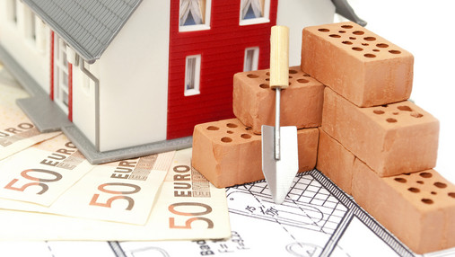 Mission Eigenheim - planen, finanzieren, absichern Foto zum Thema "Immobilienfinanzierung": Zu sehen ist ein Modellhaus, das 50-Euro-Banknoten steht. Davor Modell-Backsteine und eine kleine Schaufel.